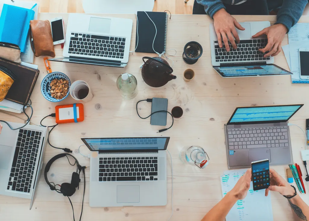 Web design team collaborating at desk with laptops, snacks and various tech accessories.