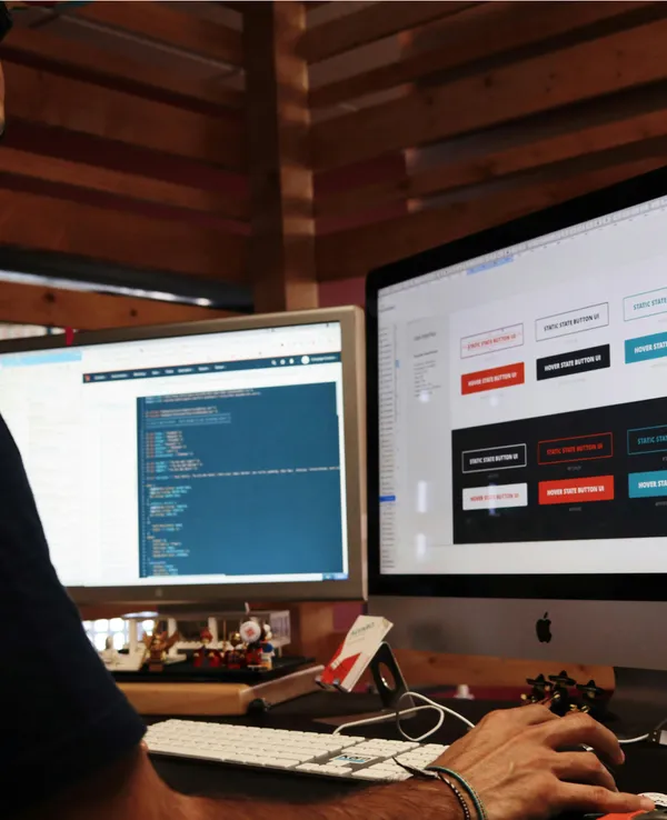 Web designer crafts responsive layout on dual monitors, showcasing code and button designs.